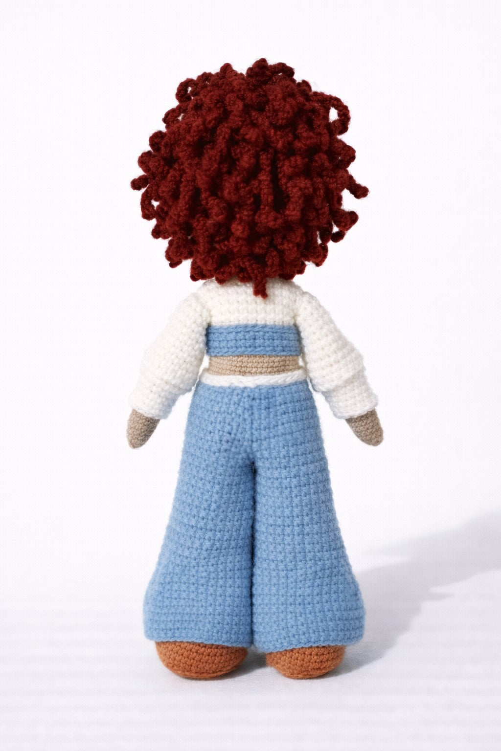 Crocheted doll with red hair, white sweater, and blue pants on a white background