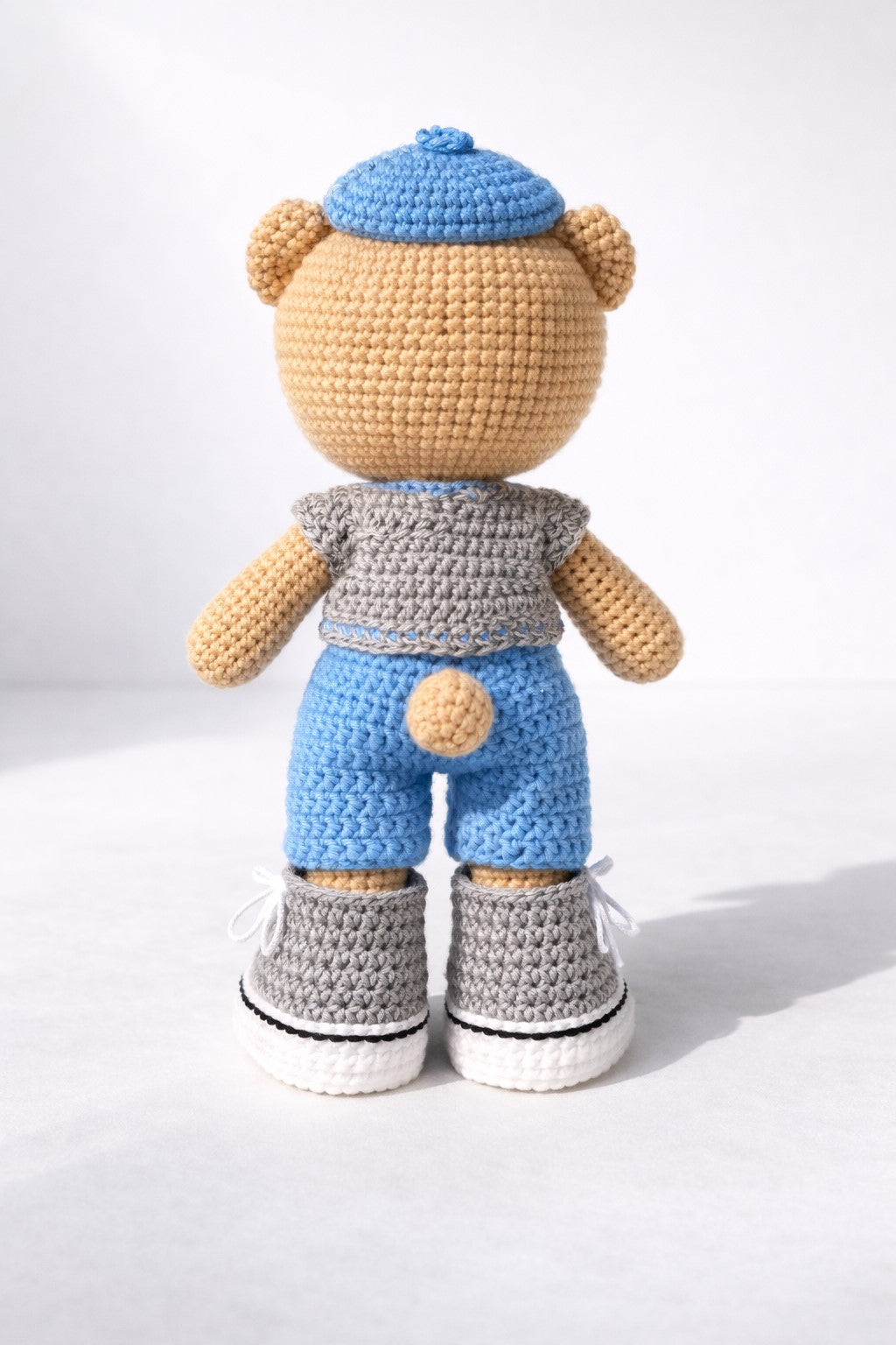 Crocheted teddy bear with blue hat, gray shirt, and pants on a white background