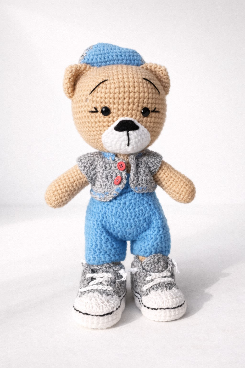 Crocheted teddy bear wearing a blue hat, gray shirt, blue overalls, and gray shoes on a white background