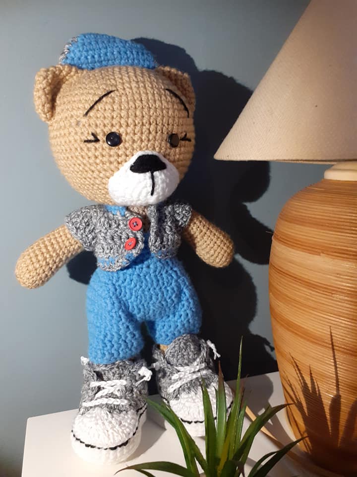 Crocheted teddy bear doll with a blue hat and shoes on a surface next to a lamp.