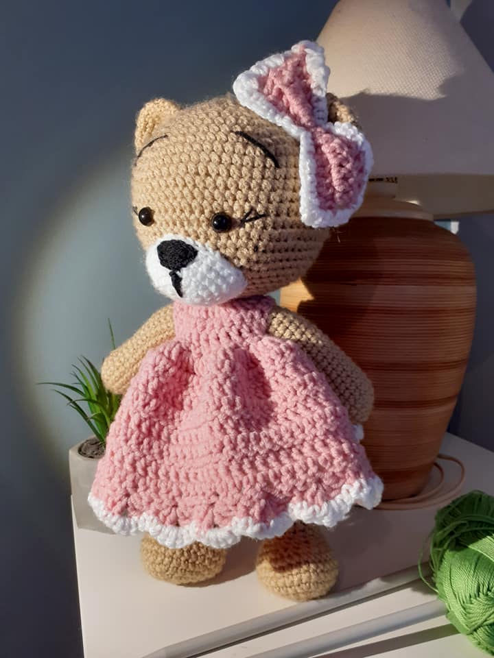 Crocheted teddy bear in a pink dress with a white bow on a neutral background
