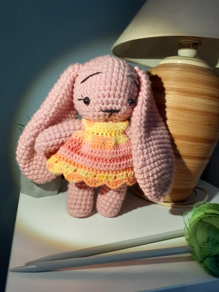 Pink crochet bunny with a yellow dress on a table next to a lamp and pen.