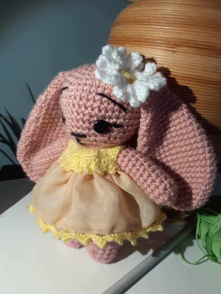 crocheted bunny doll with a flower on its head and a dress, sitting on a surface.