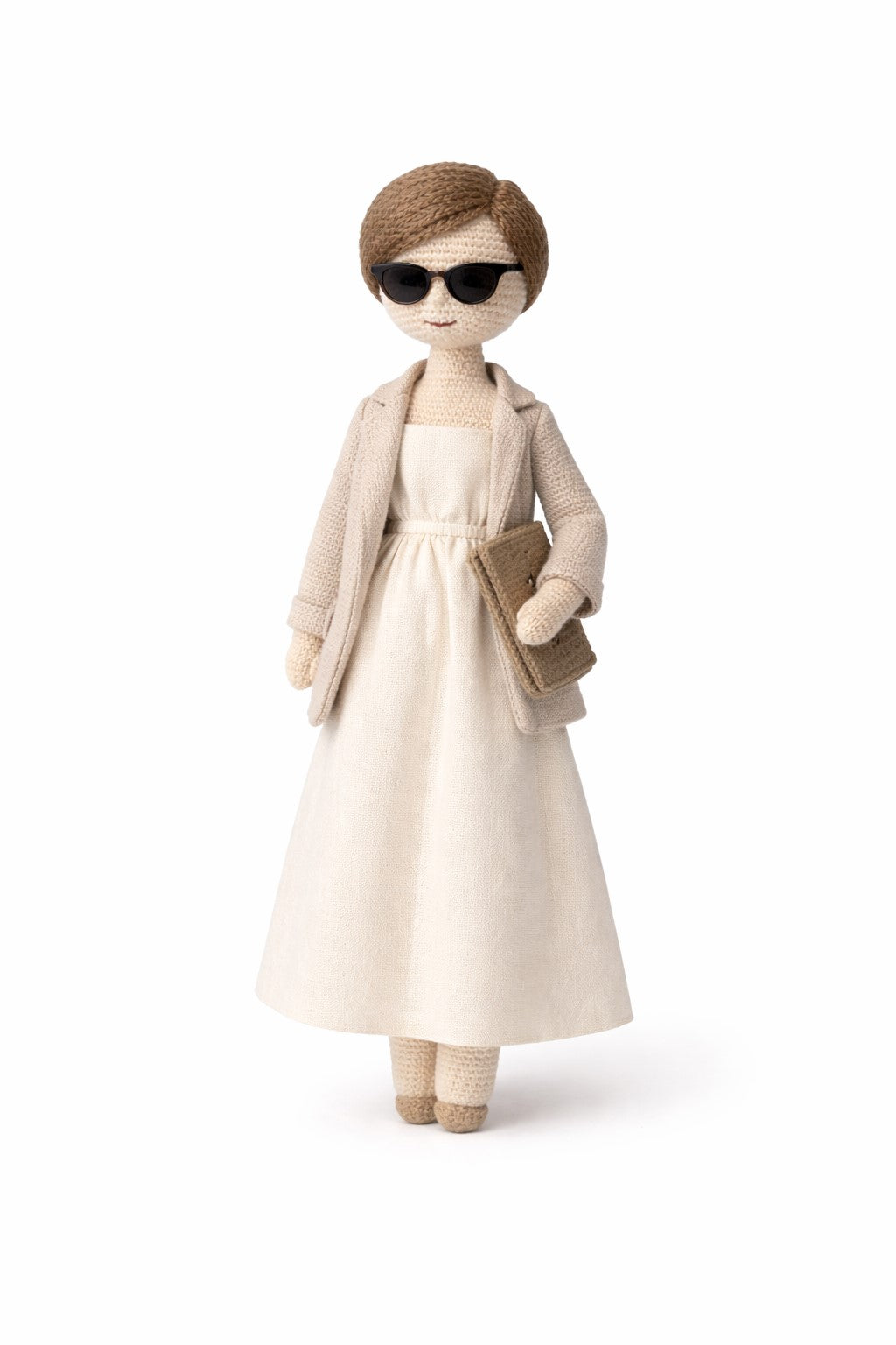 Crocheted figure of a person wearing sunglasses, beige coat, and dress on a white background